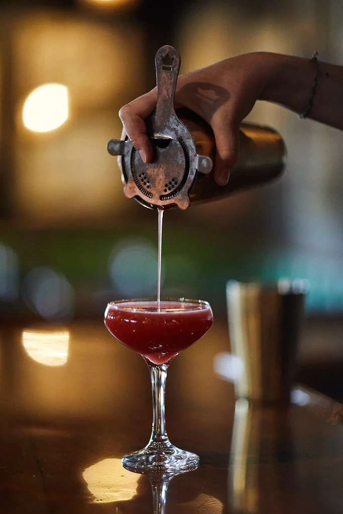 Bartender's hand pours red cocktail from shaker into coupe glass.