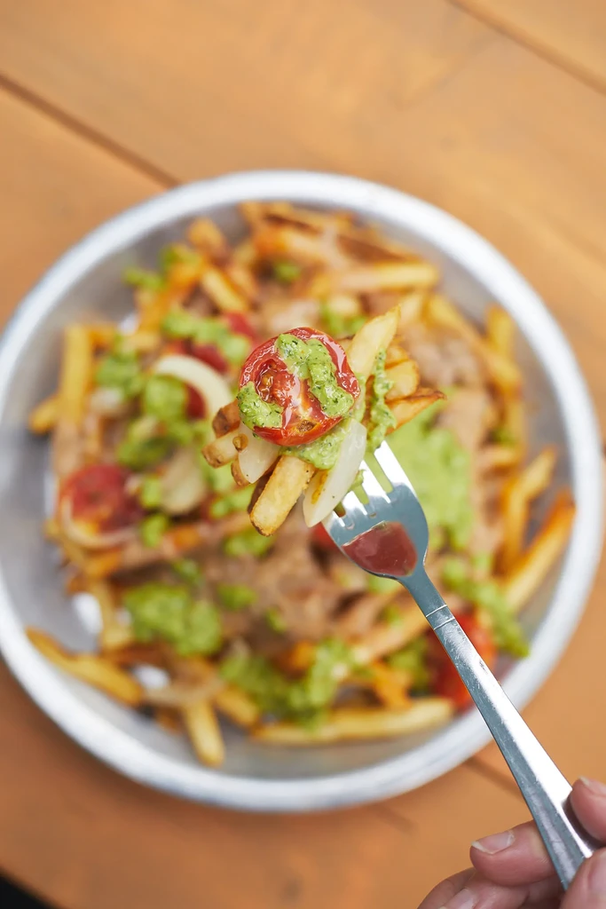 Fork lifts loaded fries topped with tomatoes, onions, and green sauce.
