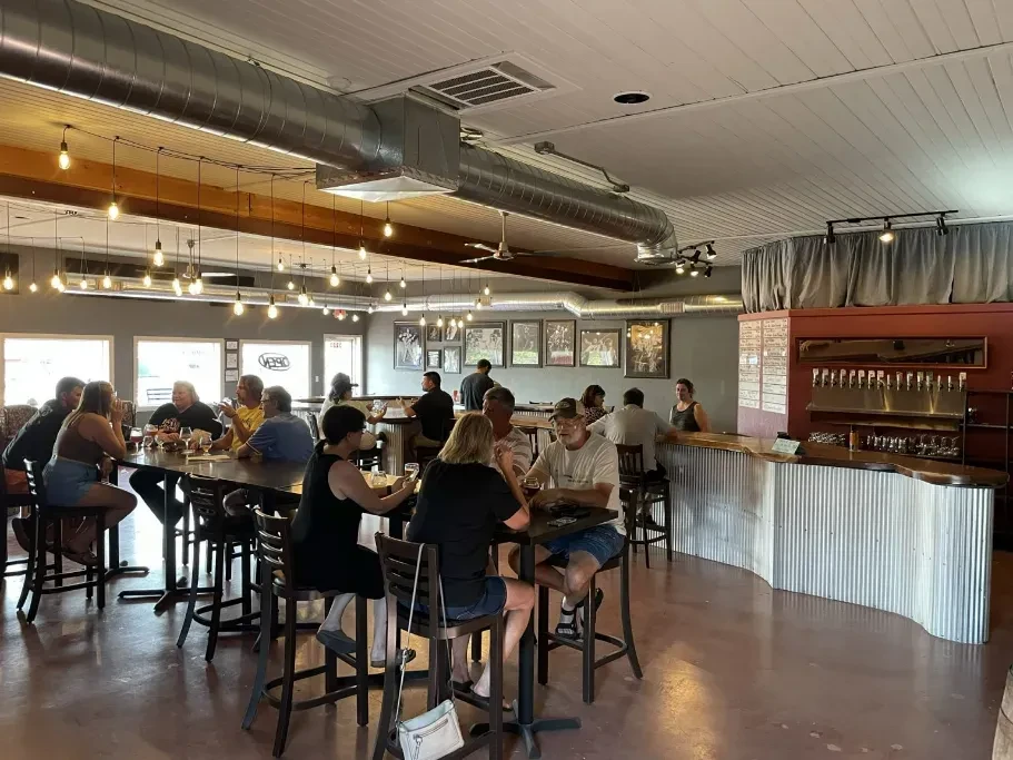 Busy brewery interior: patrons at communal table, metal bar with taps.