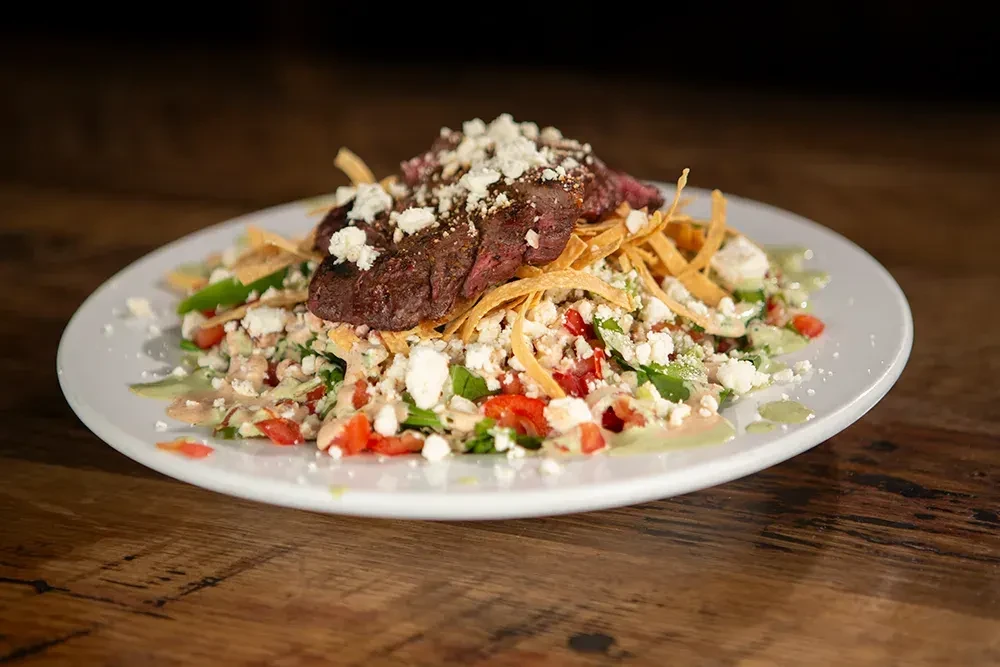 Close-up of grilled steak salad with cheese, tortilla strips, and sauces.