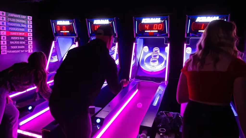 Three people play glowing Skee-Ball machines in a dim arcade.