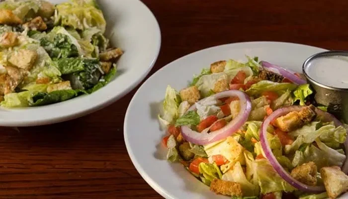 Two plates of mixed green and Caesar salads on a wooden table.