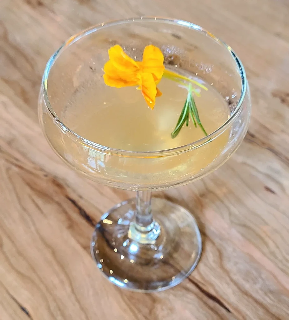Pale yellow cocktail in coupe glass with orange flower, rosemary on wood table.