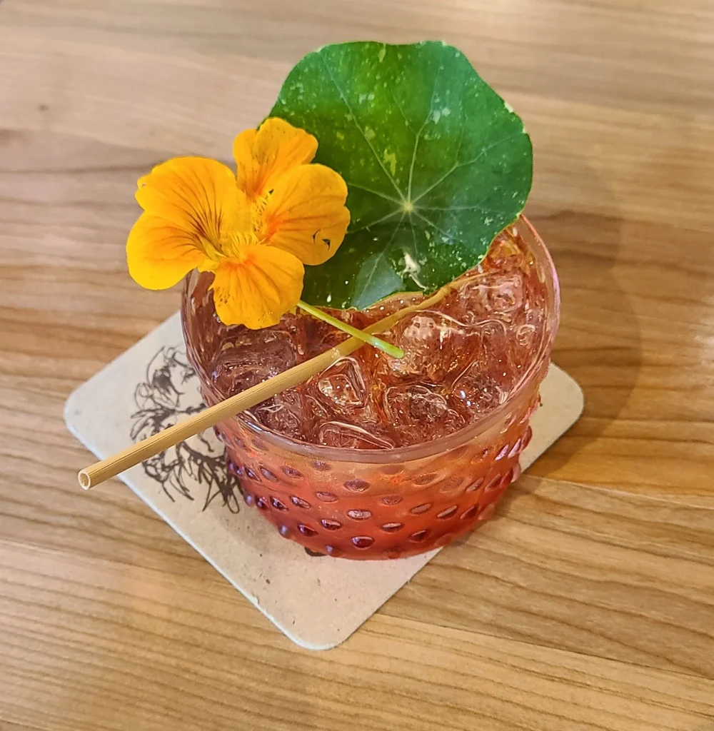 Reddish-brown cocktail with nasturtium flower and leaf on a wooden table.
