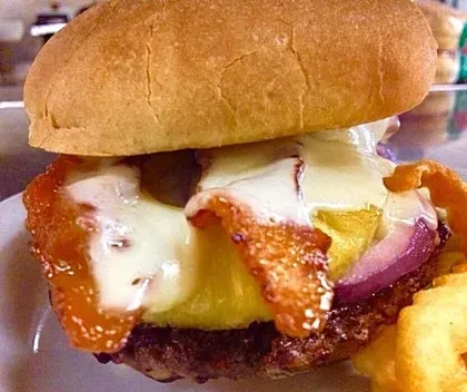 Close-up burger with melted cheese, bacon, pineapple, onion, and curly fries.