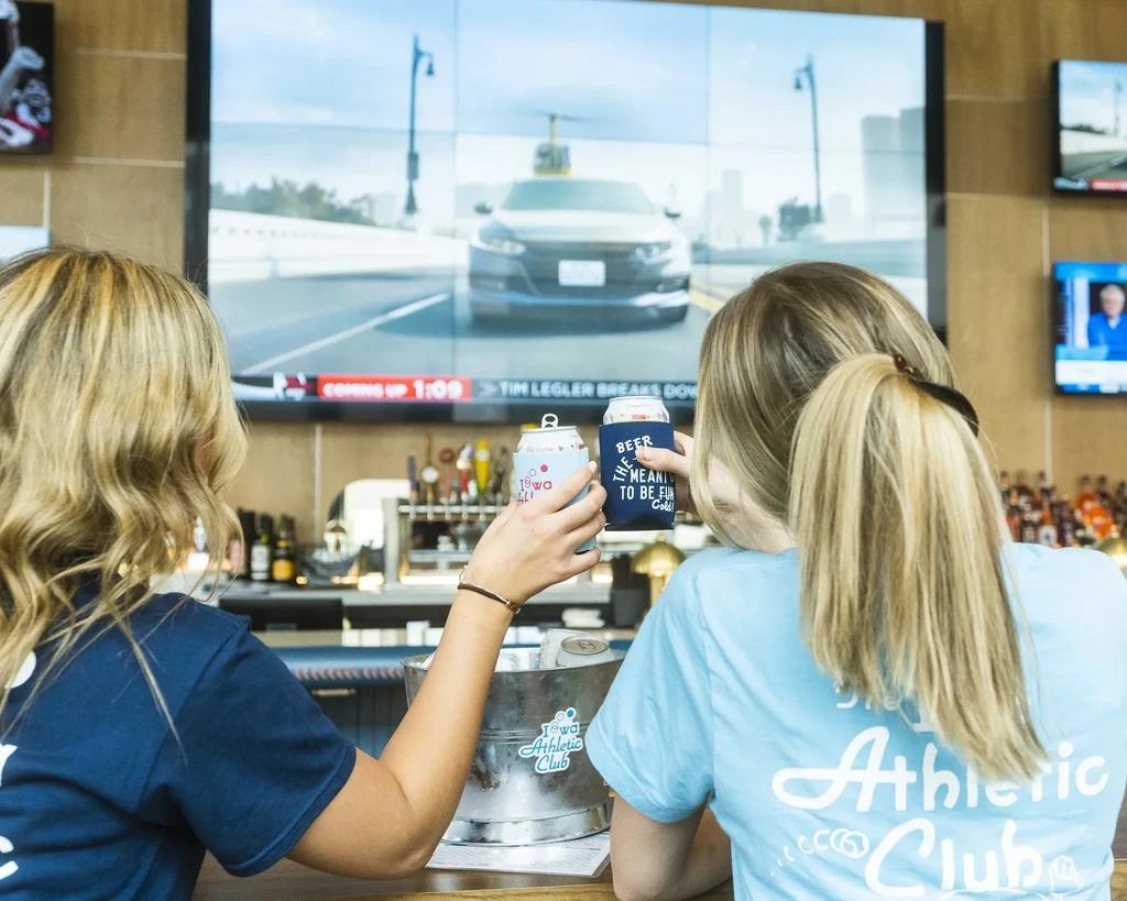 Two women drink at a bar; large video wall shows a car.