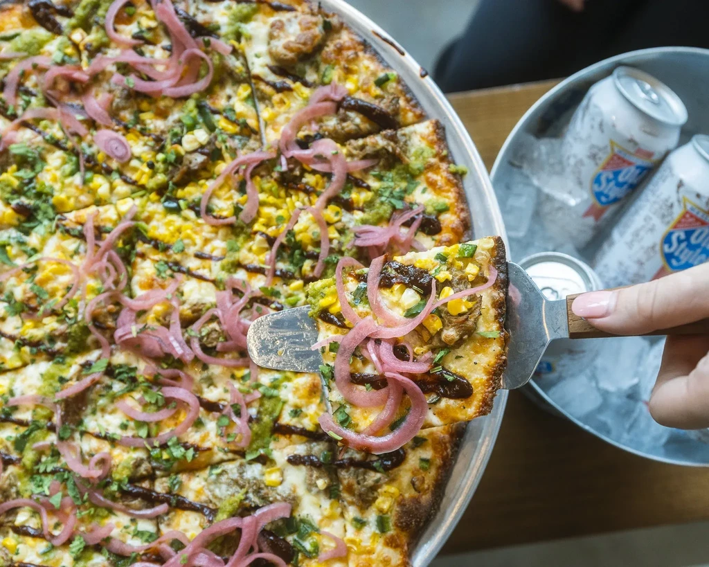 Hand lifting a pizza slice topped with corn, herbs, and red onions.