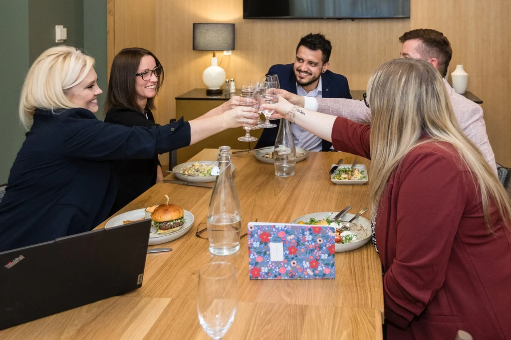 Five people toast with glasses and food at a modern restaurant.