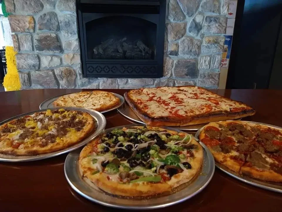 Five diverse pizzas on a dark wooden table by a stone fireplace.