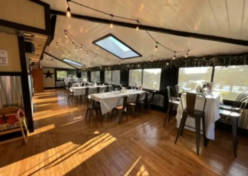 Bright, empty dining room with set tables, string lights, and water view.