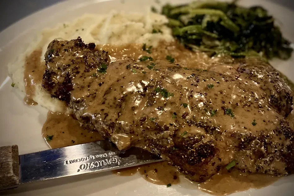 Plated steak with creamy sauce, mashed potatoes, and leafy green vegetables.