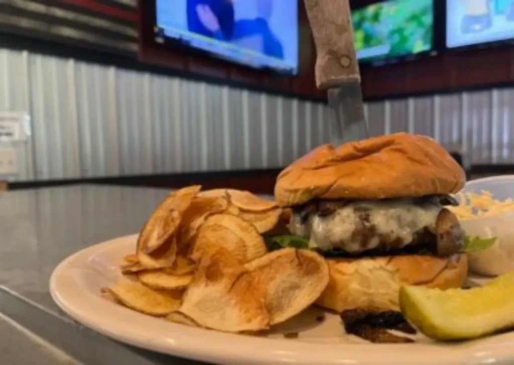 A burger held by a knife, with chips, side dish, and a pickle.