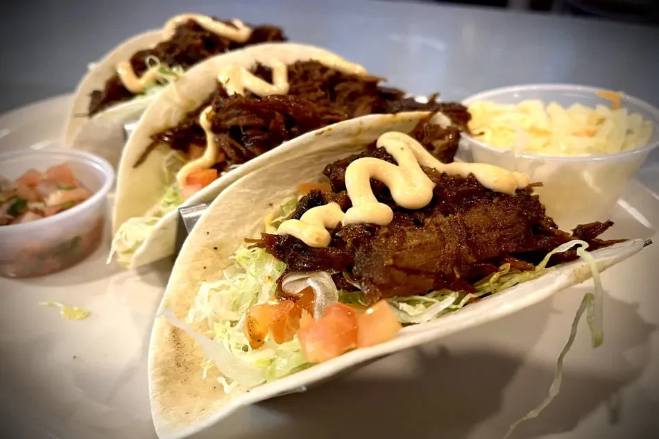 Plate of three soft-shell tacos with meat, lettuce, tomatoes, salsa, coleslaw.