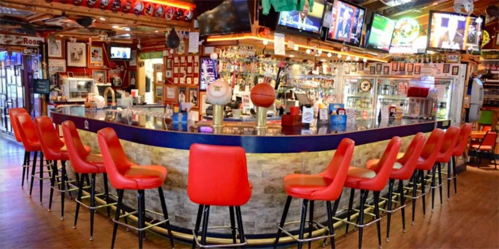 Sports-themed bar interior with curved bar, red stools, and TVs.