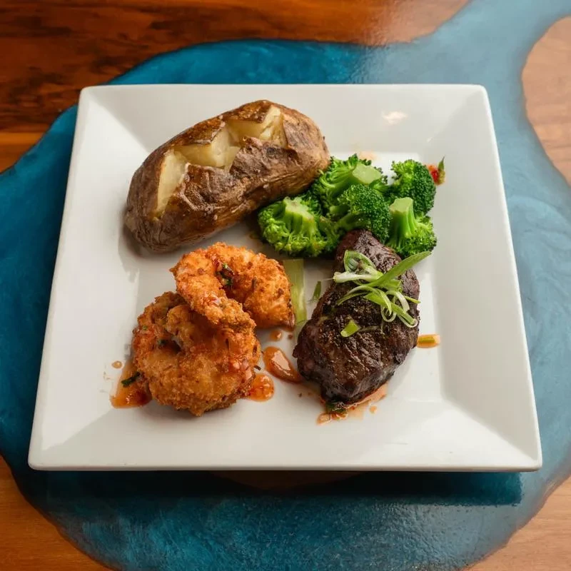 White plate with steak, shrimp, potato, broccoli on resin-inlaid wooden table.