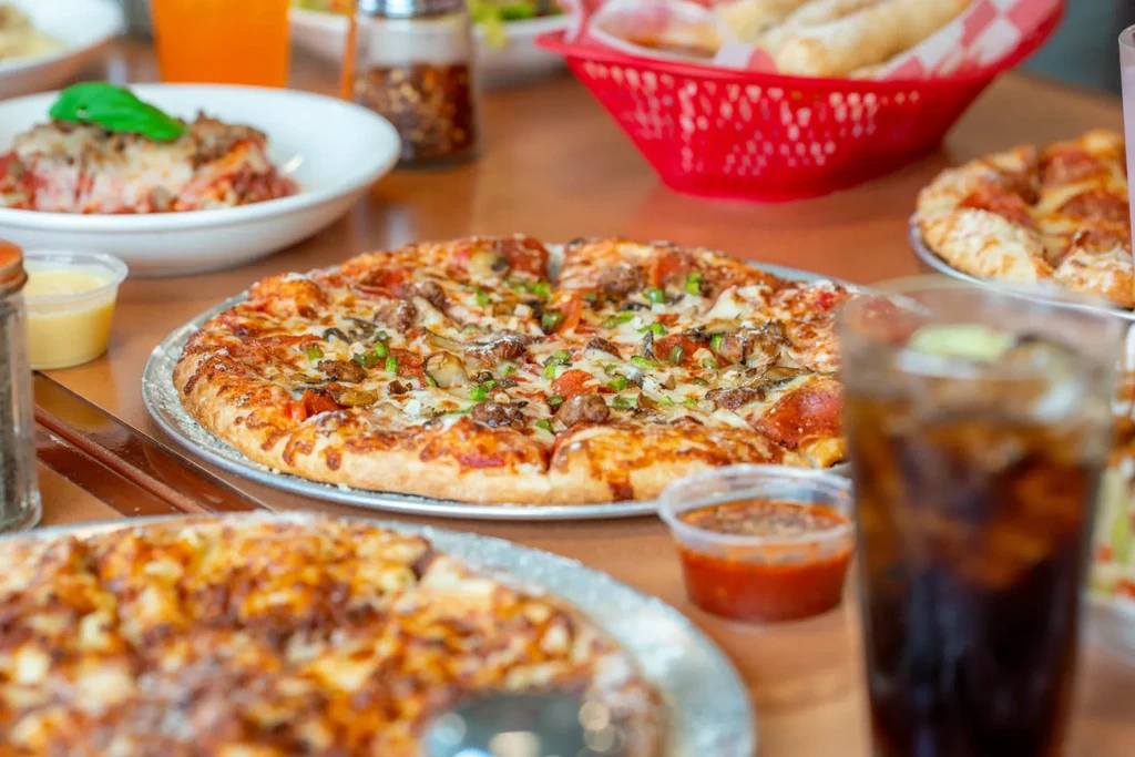 A wooden table displaying a large pizza, cheesy pasta, breadsticks, and drinks.