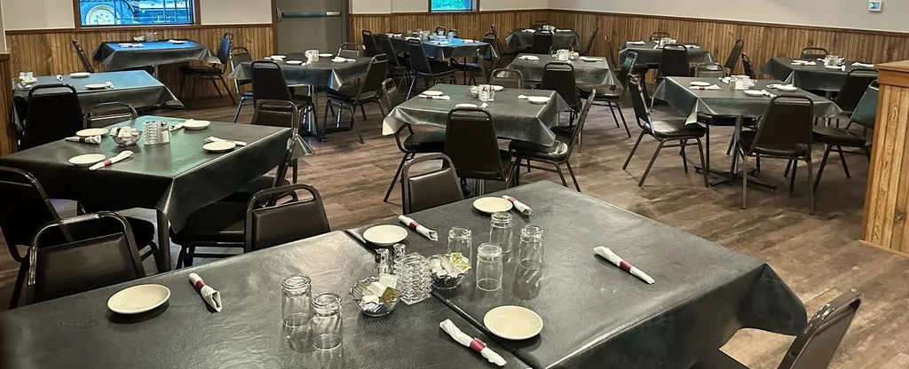 Empty casual restaurant dining room with many tables, dark cloths, white plates.