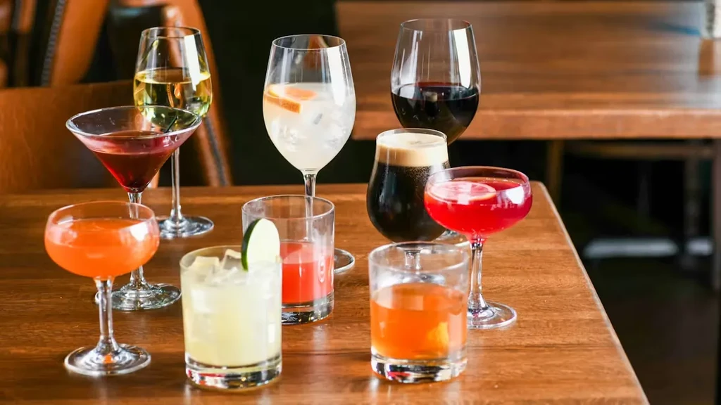 Nine garnished drinks in various glassware on a wooden table.