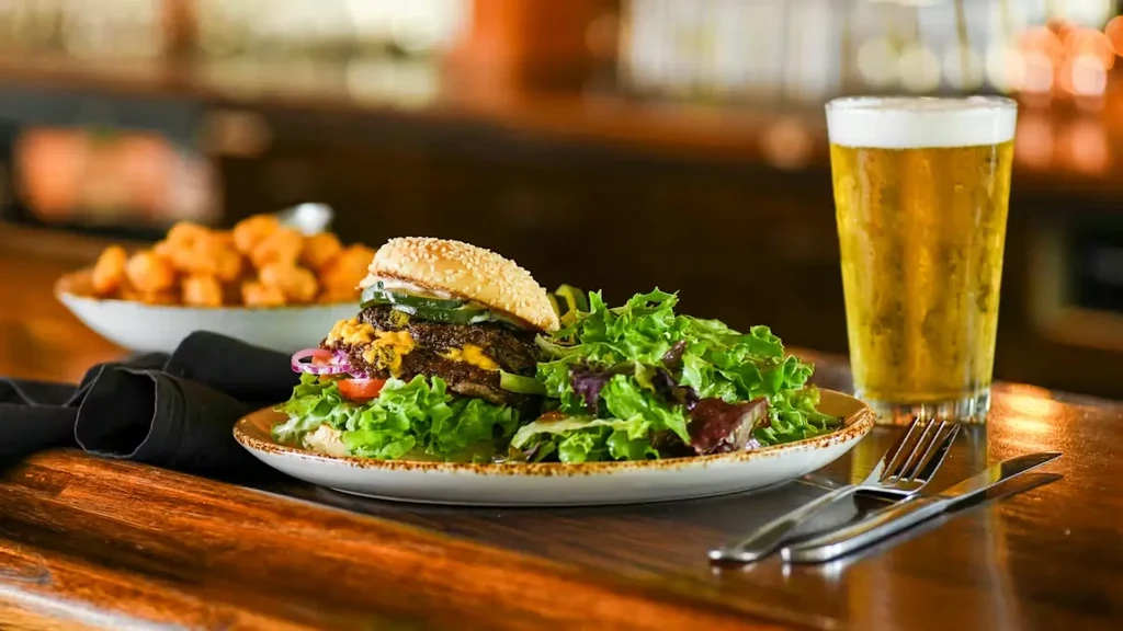 Burger, green salad, cold beer, and tater tots on a wooden table.