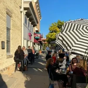 Busy sidewalk cafe with people dining under a striped umbrella.