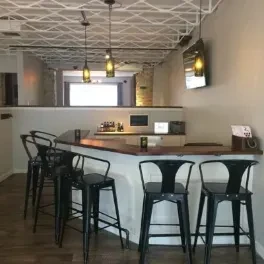 Modern bar interior featuring L-shaped counter, industrial stools, and grid ceiling.
