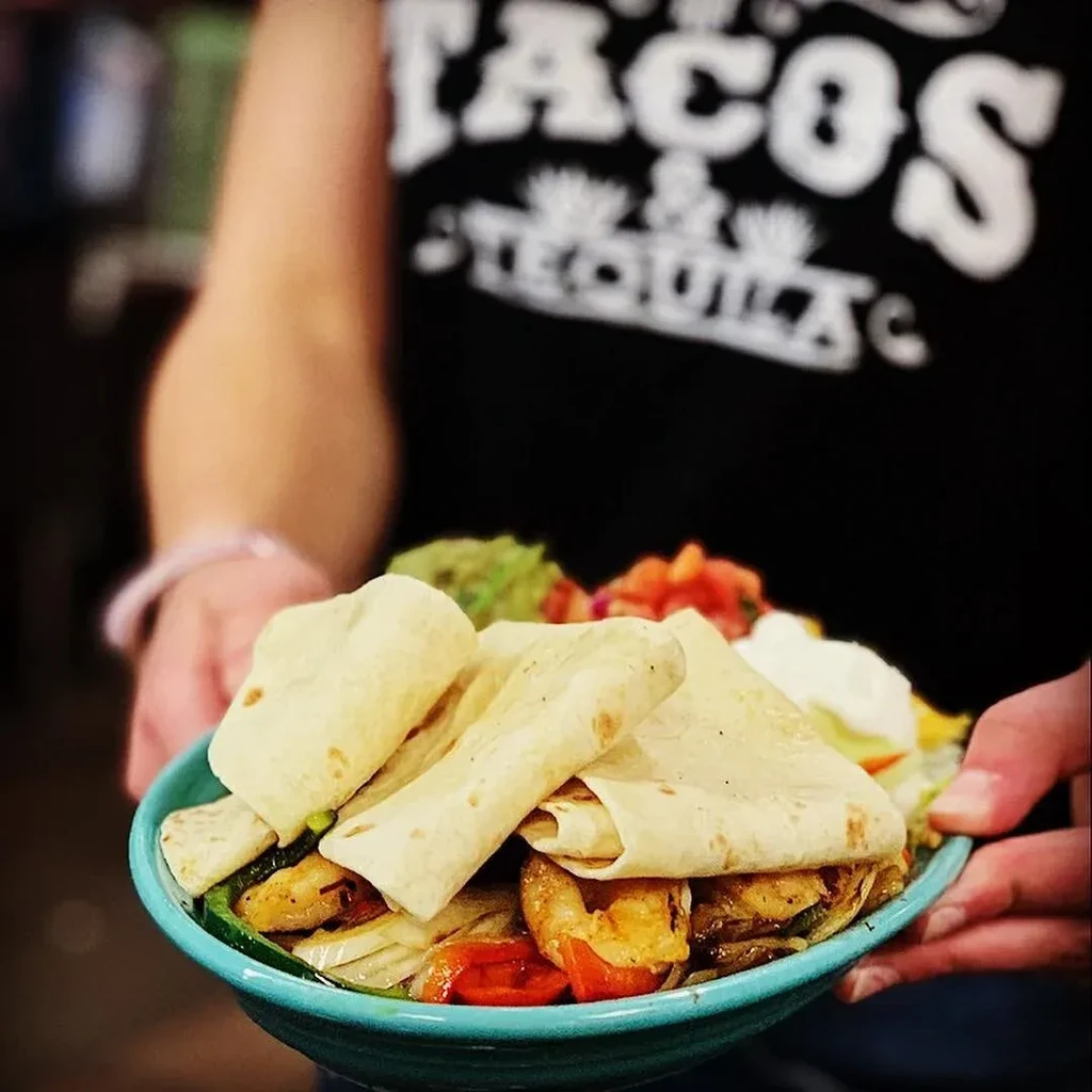 Close-up of vibrant burrito bowl held by person in "Tacos & Tequila" shirt.