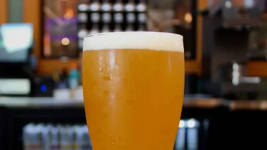 Cold glass of hazy amber beer with thick foam in a bar.