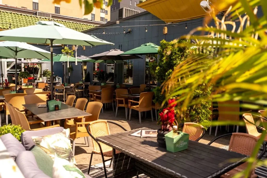 Bright outdoor restaurant patio with tables, green umbrellas, and lush plants.