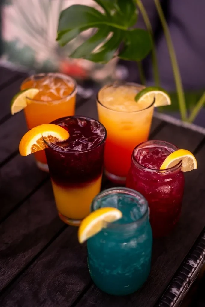 Five colorful iced drinks with fruit garnishes and a monstera leaf.