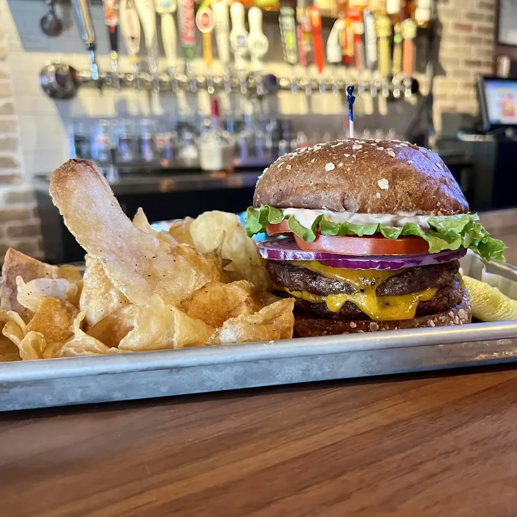 Double-patty burger, chips, pickle spear, and bar taps background.