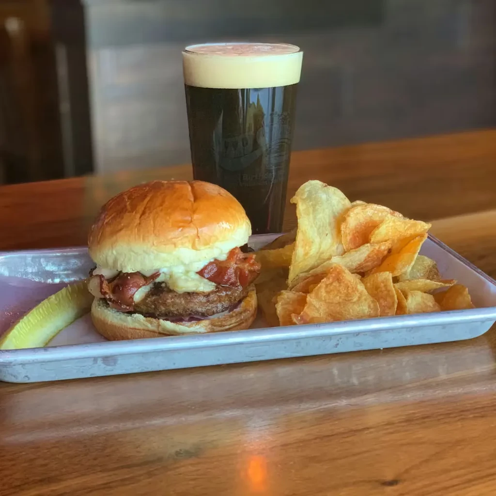 Burger with bacon, crispy chips, pickle, and dark beer on tray.