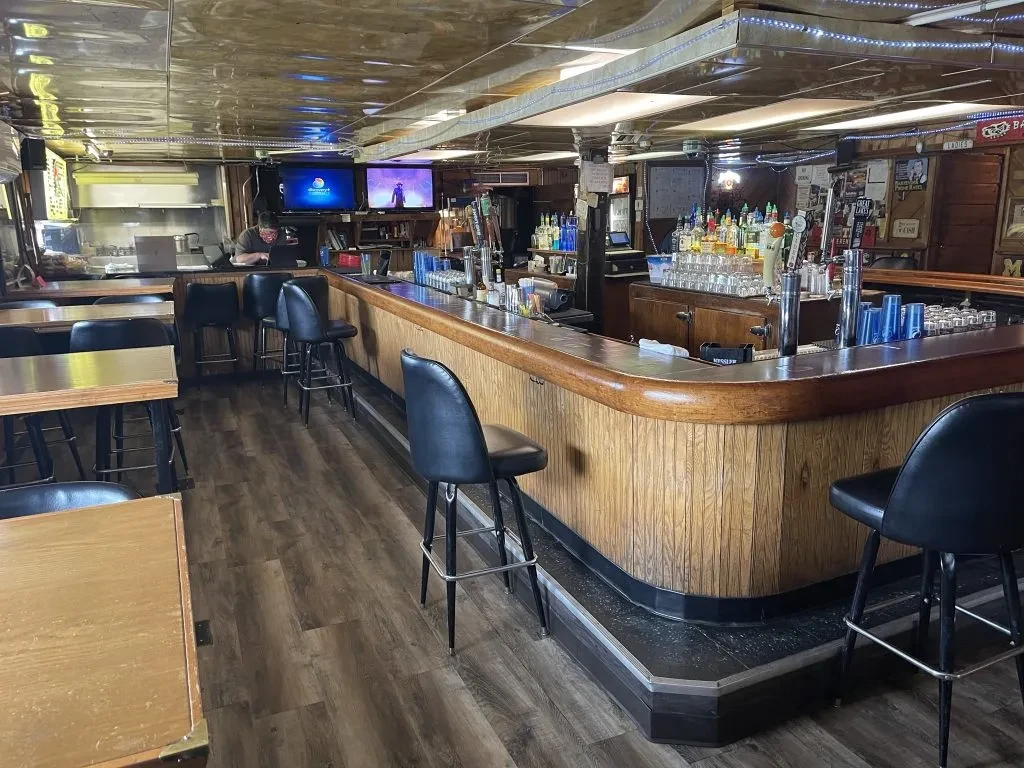 Quiet pub interior with wooden L-shaped bar, stools, liquor, and TVs.
