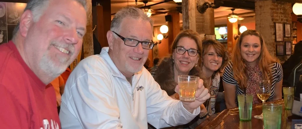 Five smiling adults at a bar with drinks in a cozy pub.