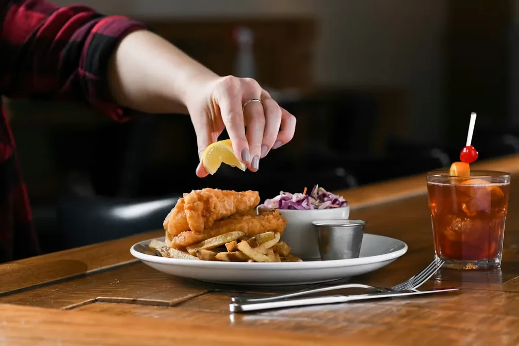 Hand squeezing lemon over fish and chips with coleslaw and a cocktail.