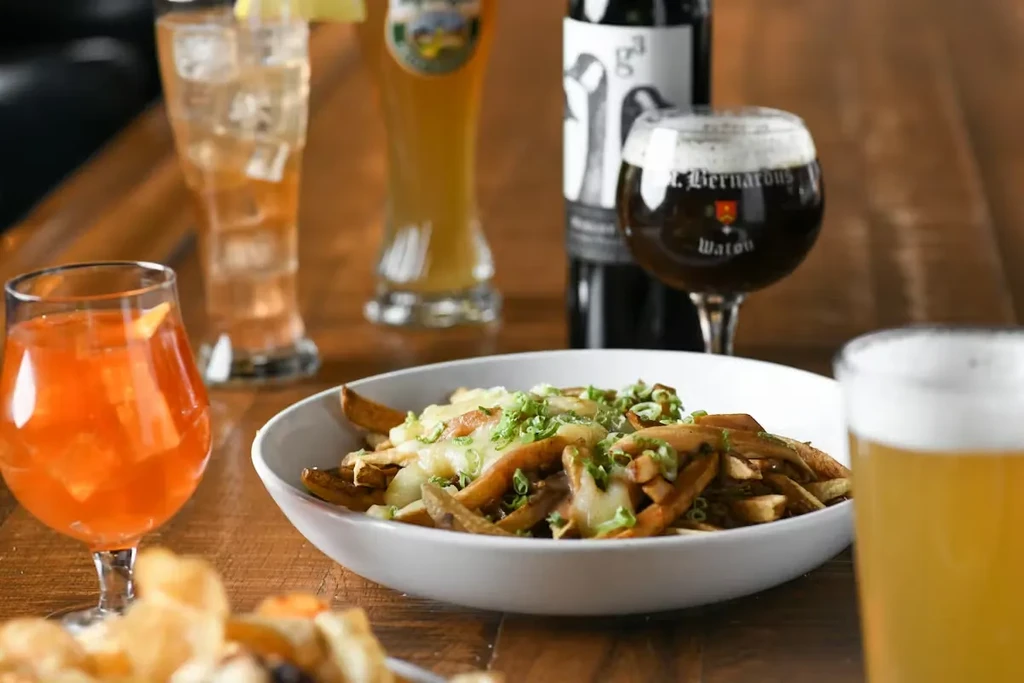 Poutine with cheese and green onions, surrounded by drinks on a wooden table.
