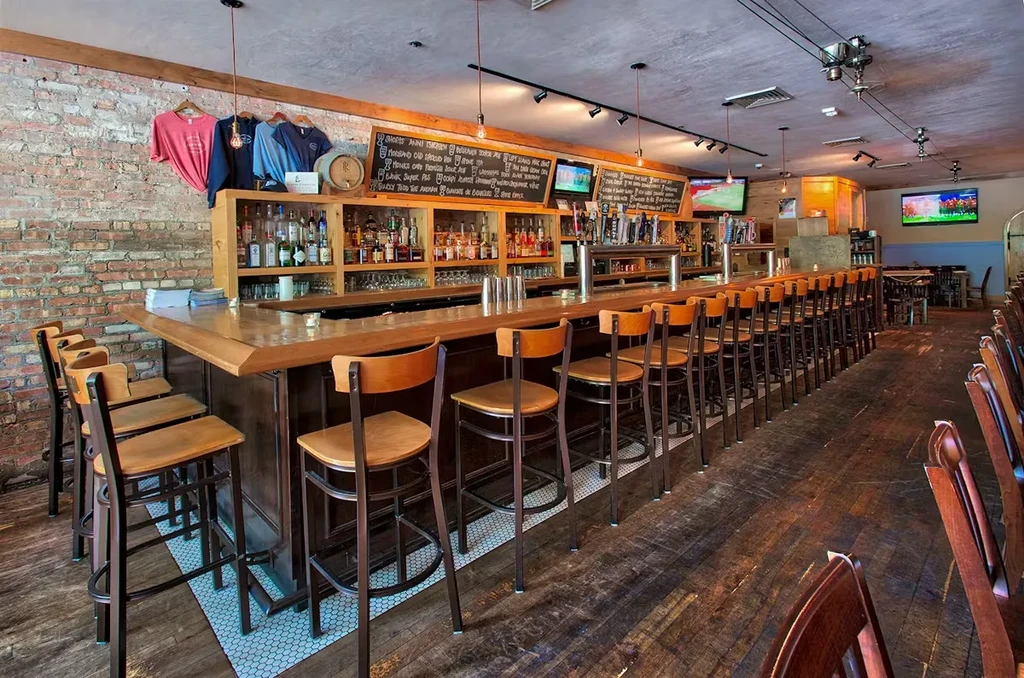 Rustic bar interior with long wooden counter, taps, and brick walls.