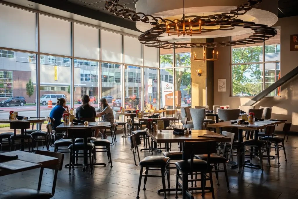 Modern restaurant interior with diverse seating, city view, and warm lighting.