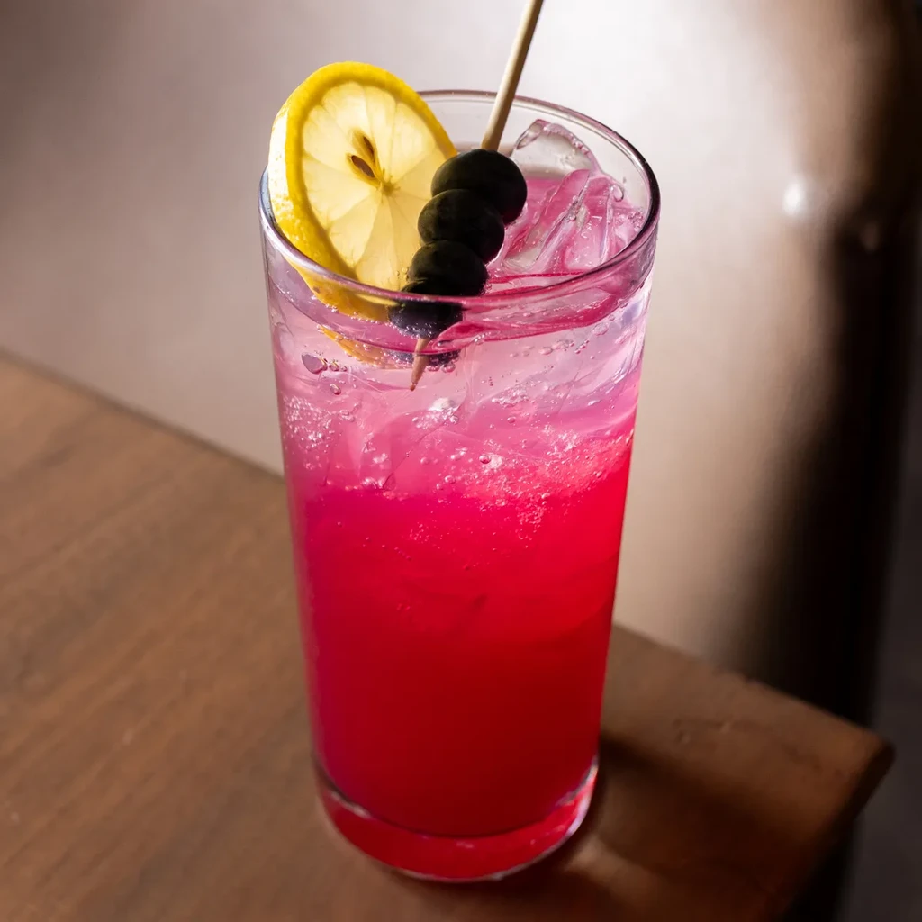 Pink carbonated drink in a glass with lemon, skewer, and ice.