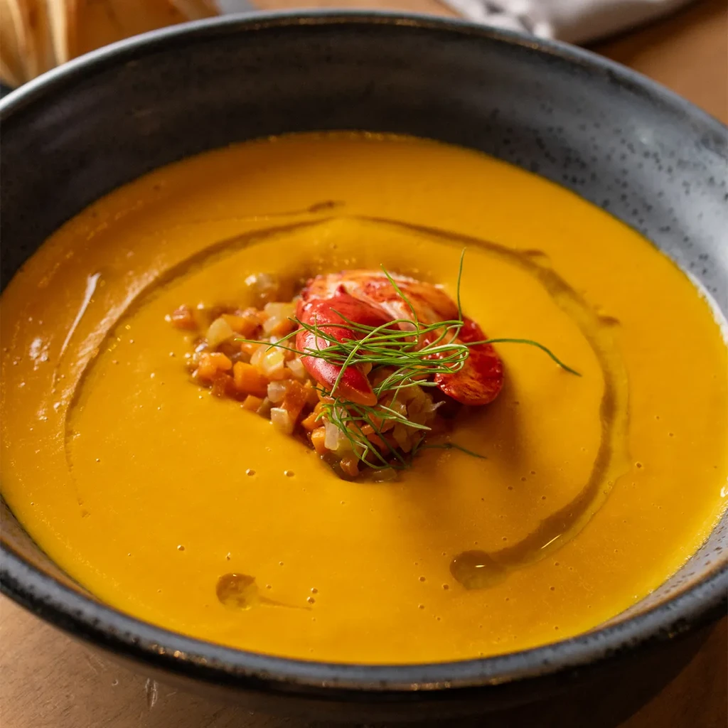 Vibrant orange-yellow bisque with lobster, vegetables, and herbs in a rustic bowl.