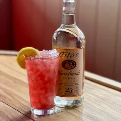 Red cocktail with lemon, Tito's vodka bottle on wooden surface.
