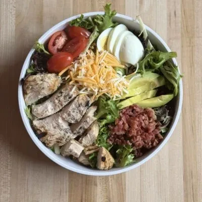 A salad with chicken, tomato, cheese, egg, avocado, bacon on lettuce.