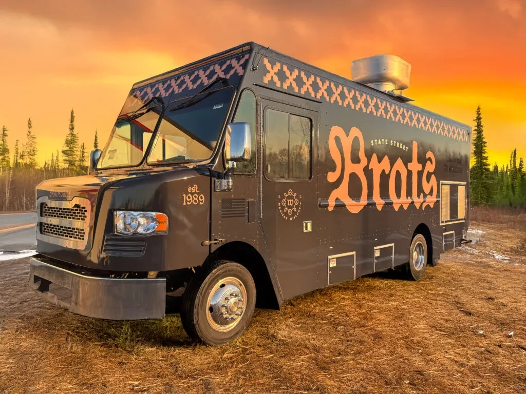 Dark gray 'STATE STREET Brats' food truck under a golden sunset sky.