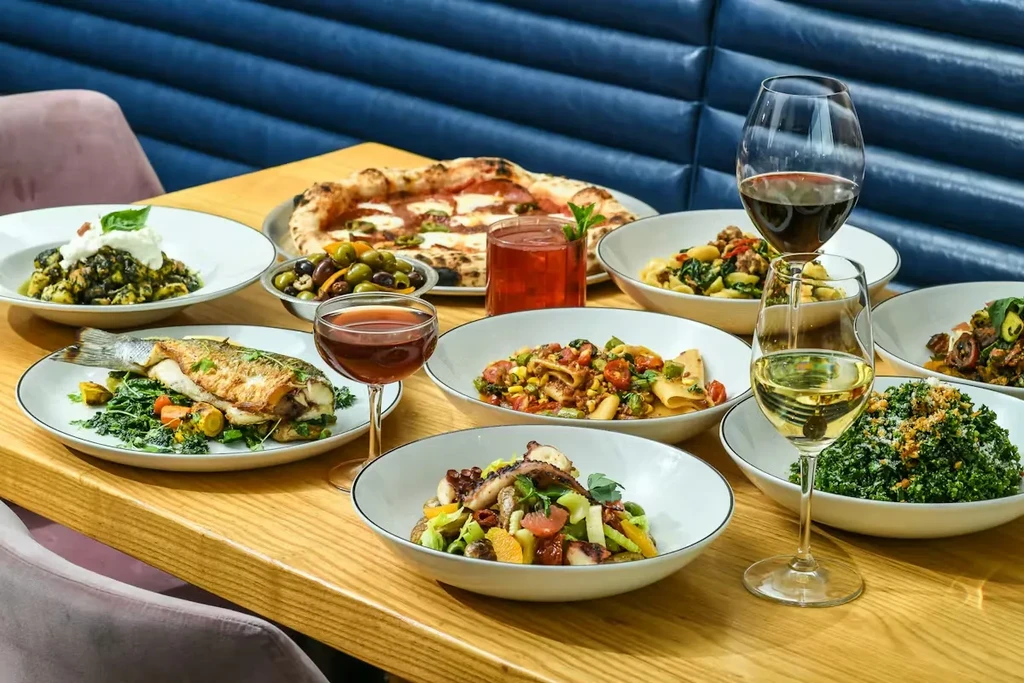 Restaurant table with pizza, roasted fish, pasta, salads, and drinks.