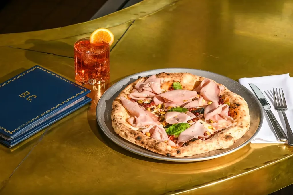 Wood-fired mortadella pizza, cocktail, and menus on a golden surface.