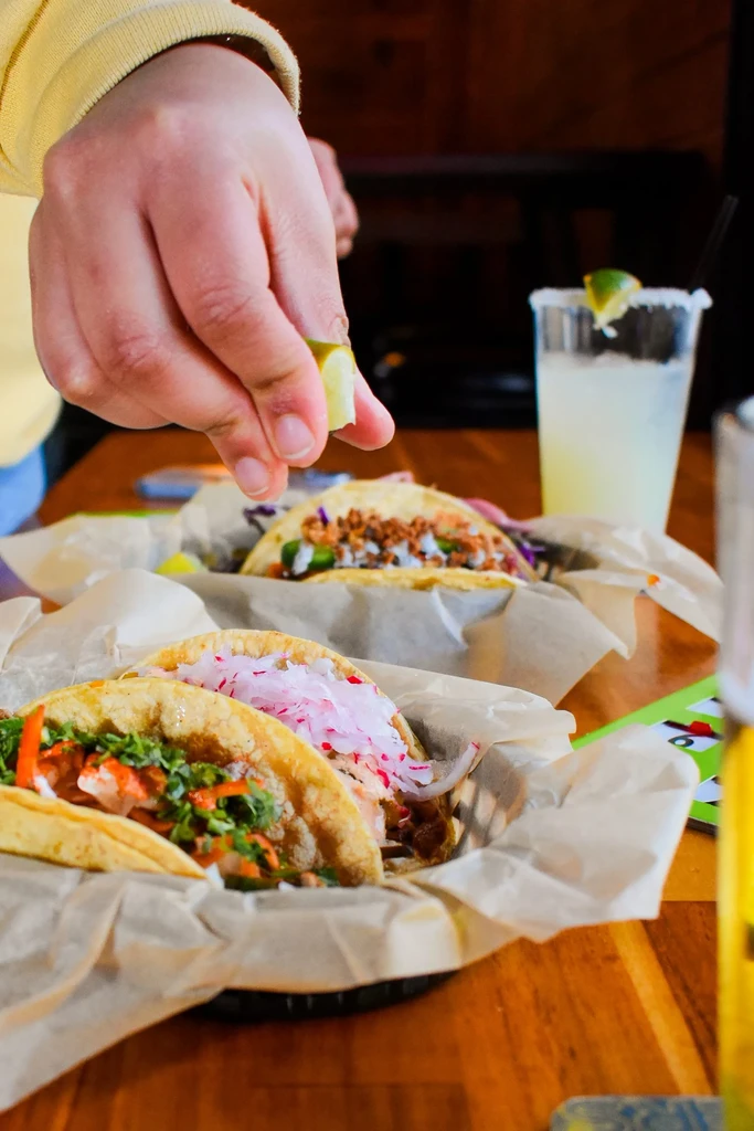 Three colorful tacos, hand squeezing lime, drink on a wooden table.