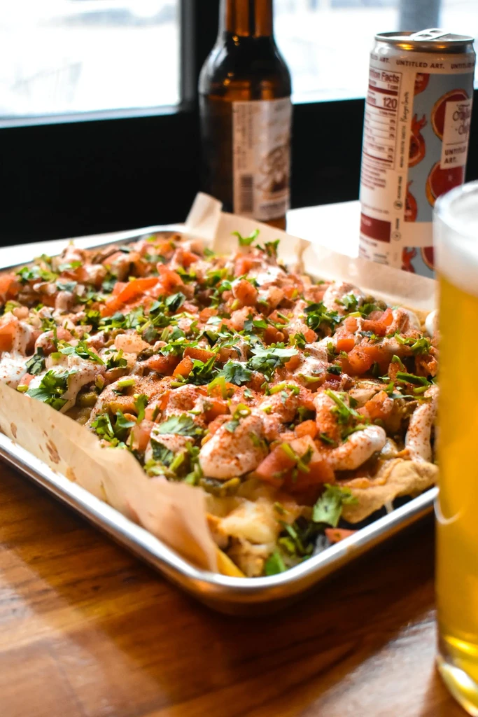 A metal tray of nachos with tomatoes, cilantro, sauce, and beer nearby.