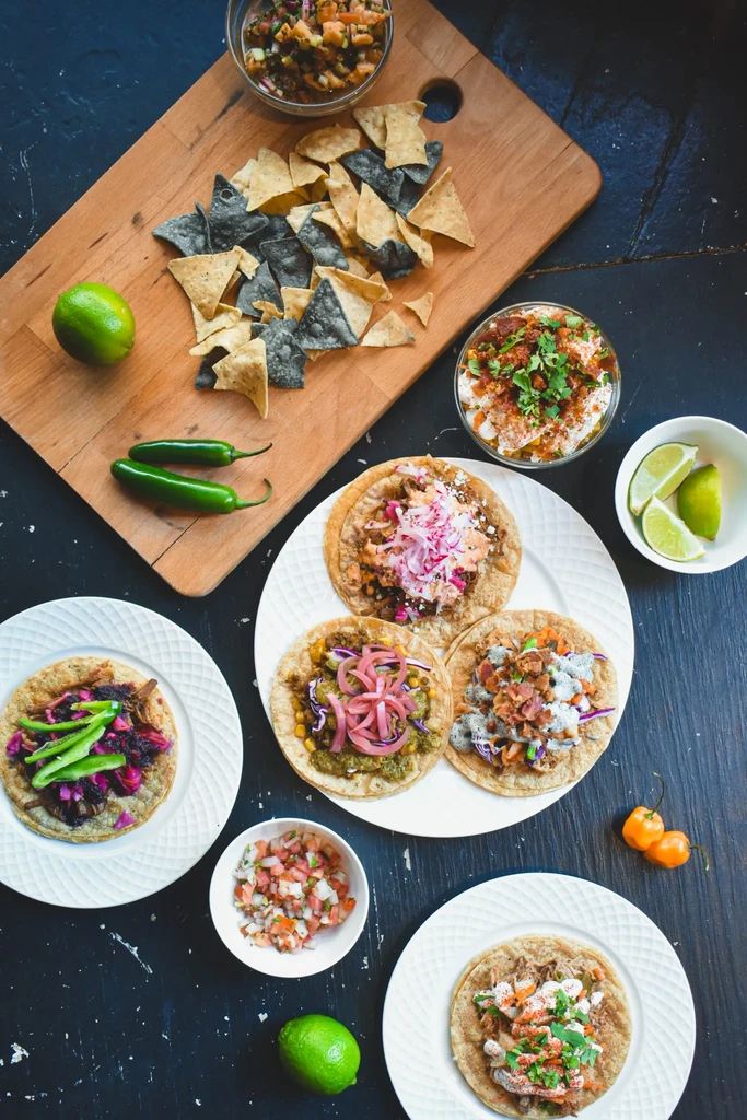 Overhead flat-lay of diverse Mexican food: tacos, chips, salsas, peppers.