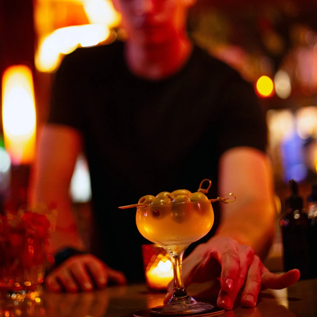 Martini with numerous olives, a hand, and lit candle on a bar.