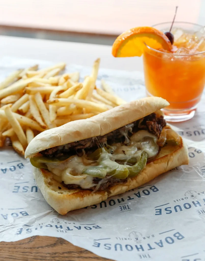 Cheesesteak sandwich, fries, and orange cocktail on a branded placemat.