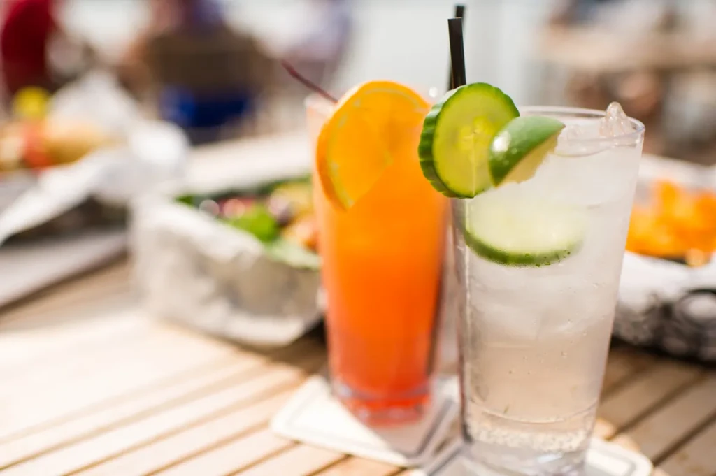 Two drinks on a wooden table: orange with orange slice, clear with cucumber.